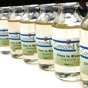 May include: A row of six clear glass bottles with blue and white labels. The labels read "Concord Soaps.com" and "Lilacs in Bloom Fragrance Oil, Weight: 1 oz."