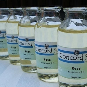 May include: Six clear glass bottles with a light yellow liquid inside. Each bottle has a white label with black text that reads "Concord Soap Co." and "Rose Fragrance Oil" with a weight of 1 oz. The bottles are lined up in a row on a white and gray striped surface.