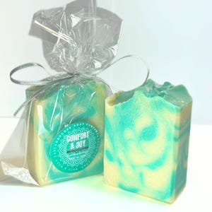 May include: Two handmade soaps with a swirl design in teal and cream. One soap is wrapped in clear cellophane and tied with a silver ribbon. The wrapped soap has a teal sticker that reads "Comfort & Joy".