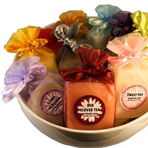 May include: A white bowl containing various handmade soaps, each encased in coloured tulle and tied with ribbons. Soaps are labelled "Lilacs in Bloom", "Red Clover Tea", and "Sweet Pea". The soaps are from The Concord Soap Company.