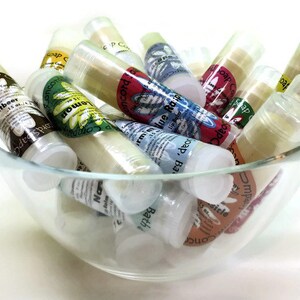 May include: A glass bowl filled with various small, clear tubes containing lip balm. The tubes have colorful labels with different scents and flavors, including "Rootbeer", "Lemon", "Blue Raspberry", "Bath", "Vanilla", and "Impish Berry".