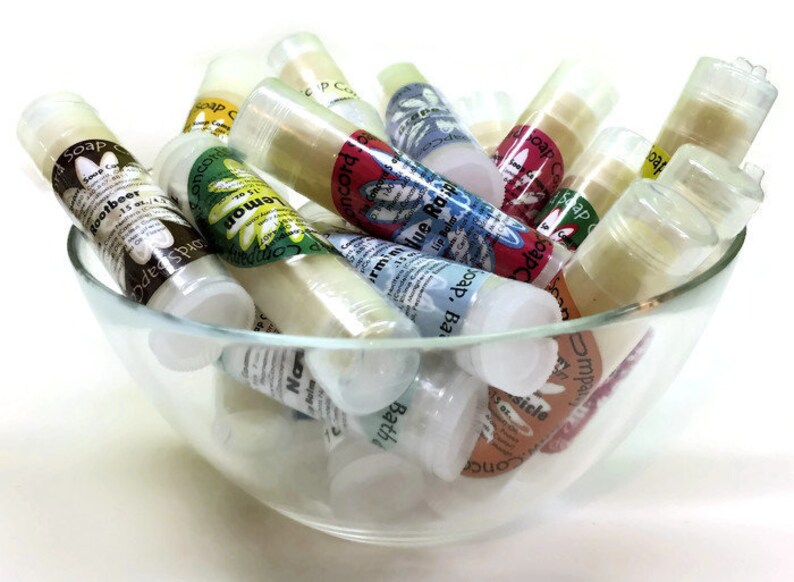May include: A glass bowl filled with various small, clear tubes containing lip balm. The tubes have colorful labels with different scents and flavors, including "Rootbeer", "Lemon", "Blue Raspberry", "Bath", "Vanilla", and "Impish Berry".