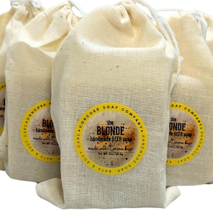 May include: Four small white cloth bags with drawstring closures, each containing a bar of handmade beer soap. The soap is labeled "The Blonde" and is made with Corona beer. The bags are arranged in a row, with the soap visible in the front bag.