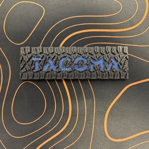 May include: A black rubber patch with the word "TACOMA" embossed in blue letters. The patch has a tire tread pattern.