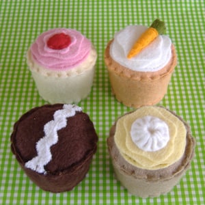 Puede incluir: Cuatro cupcakes de fieltro con diferentes diseños de glaseado. Un cupcake tiene glaseado rosa con un diseño de remolino, uno tiene glaseado blanco con una zanahoria en la parte superior, uno tiene glaseado blanco con un diseño de remolino y uno tiene glaseado marrón con un diseño de remolino blanco.