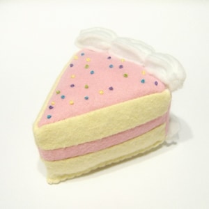 Pode incluir: Uma fatia de bolo de feltro rosa e amarelo com cobertura branca e granulado colorido. O bolo é feito à mão e tem uma aparência tridimensional.