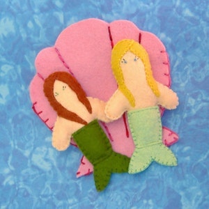 May include: Two felt mermaids, one with brown hair and a green tail, the other with blonde hair and a light blue tail, are sitting inside a pink felt seashell.