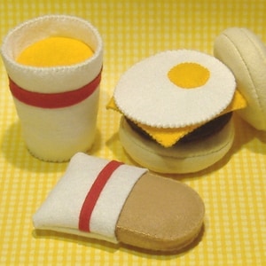 May include: Felt food play set with a white cup with a red stripe, a white and yellow egg sandwich, and a brown and white felt hot dog in a white wrapper with a red stripe.