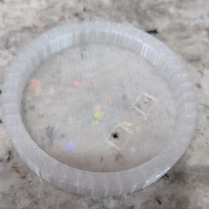 May include: A circular, translucent white tray with a ribbed outer edge and a flat base. The surface features embedded iridescent, multi-colored foil flakes that catch the light, along with two small, square-shaped cutouts near the center.