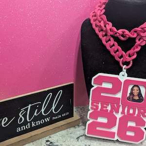 May include: A pink chain necklace with a large pendant featuring the text "20 SENIOR 26" and a photo. The necklace is displayed on a black mannequin head against a pink background. A small sign reads "be still and know Psalm 46:10".