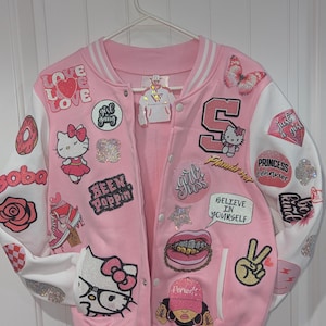 May include: Pink and white varsity jacket with multiple patches. Patches include a butterfly, Hello Kitty, the word "LOVE", and the phrase "Believe in Yourself". The jacket has a snap front closure and white sleeves.