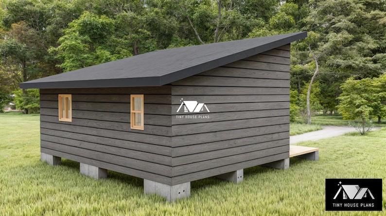 Pu&ograve; includere: Una piccola capanna con rivestimento in legno orizzontale grigio scuro, un tetto spiovente e due finestre con telai in legno chiaro. La struttura &egrave; rialzata su blocchi di cemento. Un logo con il testo TINY HOUSE PLANS &egrave; visibile sulla parete laterale.