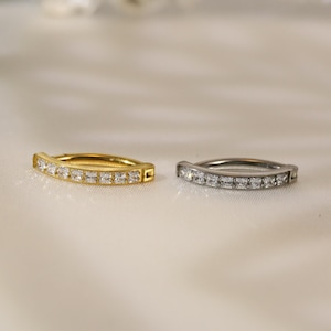 Puede incluir: Dos pendientes de aro con una fila de piedras cuadradas transparentes. El aro de la izquierda es dorado y el de la derecha es plateado.