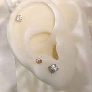May include: Three different sizes of clear cubic zirconia stud earrings. The earrings are displayed on a white ear model. The earrings are made of gold, rose gold, and silver.