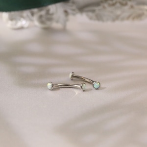 May include: Two silver curved barbell body jewelry pieces with opal-like white gemstones.