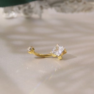 Puede incluir: Anillo de ombligo dorado con una gema cuadrada transparente y una gema redonda más pequeña.