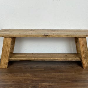 May include: A rustic wooden bench with a lower shelf. The bench is made of natural wood with a light brown finish. The bench has a simple design with straight legs and a rectangular top.