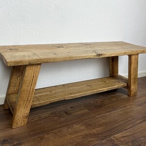 May include: A rustic wooden bench with a shelf underneath. The bench is made of natural wood with a light brown finish. The bench has a simple design with straight legs and a rectangular top.