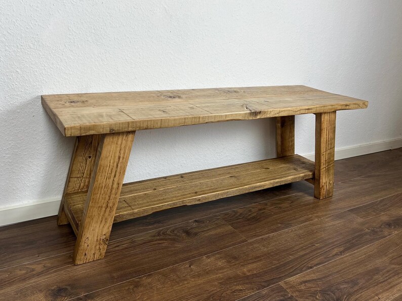 May include: A rustic wooden bench with a shelf underneath. The bench is made of natural wood with a light brown finish. The bench has a simple design with straight legs and a rectangular top.