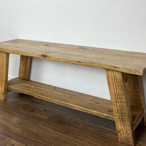 May include: A rustic wooden bench with a shelf. The bench is made of natural wood with a light brown finish. The shelf is located below the seat and is also made of wood.