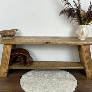 May include: A rustic wooden bench with a shelf, featuring a natural wood finish. The bench has a simple design with two angled legs on each side. The shelf is perfect for storing shoes or other items.