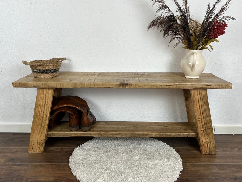 May include: A rustic wooden bench with a shelf, featuring a natural wood finish. The bench has a simple design with two angled legs on each side. The shelf is perfect for storing shoes or other items.