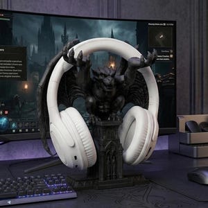 Può includere: Un paio di cuffie bianche over-ear poggiate su un supporto a forma di gargoyle grigio scuro. Il gargoyle presenta grandi ali e mani artigliate posizionate per sostenere l'archetto. La base è dettagliata con elementi architettonici gotici.