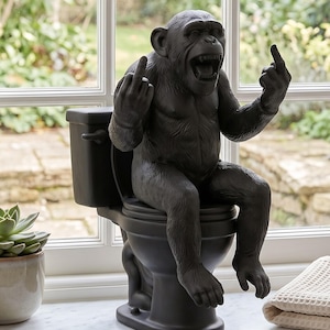Könnte beinhalten: Eine mattschwarze Figur eines Schimpansen, der auf einer Toilette sitzt. Der Schimpanse hat den Mund offen und hält beide Hände hoch, wobei die Mittelfinger in einer Geste ausgestreckt sind. Die Skulptur ist einfarbig dunkel mit Fellstruktur.