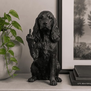 Könnte beinhalten: Eine mattschwarze Hundefigur in sitzender Position mit langen, strukturierten Ohren und detailliertem Fell. Der Hund hebt seine rechte Vorderpfote mit ausgestrecktem Mittelfinger in einer Geste. Die Skulptur weist realistische Gesichtszüge auf.