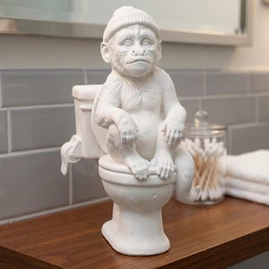Könnte beinhalten: Eine mattweiße Skulptur eines Affen mit Mütze, der auf einer Miniaturtoilette sitzt. Der Affe weist eine detaillierte Fellstruktur und einen neutralen Gesichtsausdruck auf. Die Skulptur ist ein einteiliges Objekt, das den Affen auf dem Sitz zeigt.
