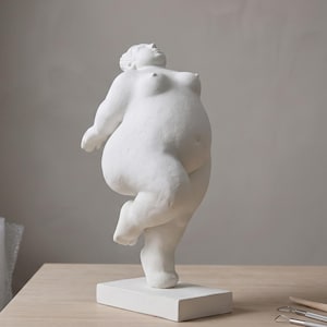 Op de afbeelding: Een wit mat beeldhouwwerk van een volslank persoon in een dynamische danshouding met één been opgetild. Het figuur heeft een glad, getextureerd oppervlak en staat op een rechthoekige voet, wat een gevoel van beweging uitstraalt.
