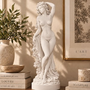 May include: A white matte sculpture of a standing figure with long wavy hair adorned with flowers, one arm raised above the head, and draped fabric cascading down the side. The figure stands on a circular base with a smooth, neutral finish.
