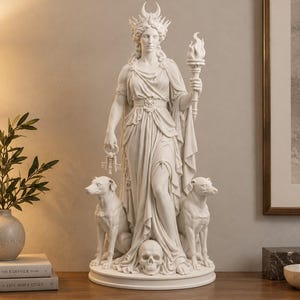 Może przedstawiać: Biała statua przedstawiająca stojącą postać w koronie z półksiężycem, trzymającą w jednej dłoni płonącą pochodnię, a w drugiej pęk kluczy. U stóp postaci siedzą dwa psy, otaczające ludzką czaszkę umieszczoną na okrągłej podstawie rzeźby.