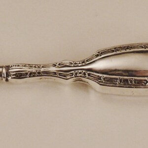 May include: A close-up of a silver utensil with an ornate handle and a rounded, elongated head. The handle features detailed decorative patterns and a series of small, rounded sections. The head has a smooth, reflective surface with a decorative border.