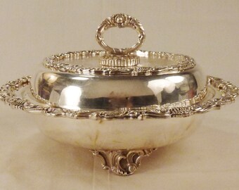 May include: A silver-colored, ornate butter dish with a domed lid and a decorative handle. The dish has a scalloped edge and sits on three ornate feet. The lid and base are highly reflective, suggesting a polished finish.