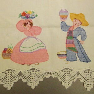 Op de afbeelding: Geborduurd wit linnen textiel met twee figuren. Eén draagt bloemen, de ander een vaas. De onderrand heeft een decoratieve witte kanten rand. De figuren dragen kleurrijke kleding.