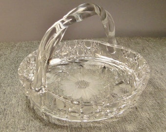 May include: A clear, oval-shaped crystal basket with a twisted handle. The basket features a floral design on the base and a cut-glass pattern around the rim. The handle is securely attached to the sides of the basket.