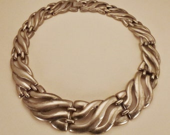 May include: A silver-toned necklace with a unique, wave-like design. The necklace features a series of interconnected, curved links that create a flowing, textured appearance. The clasp is a simple, secure closure. This statement piece is perfect for adding a touch of elegance.