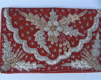 Könnte beinhalten: Eine rechteckige, rote Clutch mit aufwendiger Silber- und Goldperlenstickerei. Das Design zeigt Blumen- und Blattmotive sowie sternförmige Akzente. Die Kanten sind ebenfalls mit Perlen besetzt, wodurch ein dekorativer Rand entsteht.