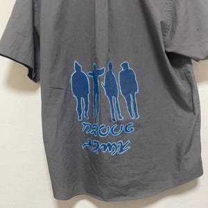 May include: Gray short-sleeved button-down shirt with a blue silhouette of five people and the text "DRUG ARMY" printed on the back.