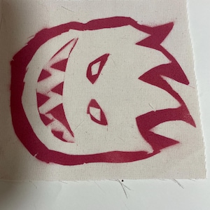 May include: A red, hand-painted monster face on a white canvas fabric. The monster has a wide, toothy grin and two small eyes.