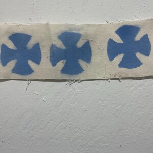 May include: A strip of off-white fabric featuring three repeating blue cross designs. The crosses have a stylized, four-armed shape. The fabric has a slightly frayed edge, and the background is a plain white wall.