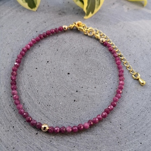 Könnte beinhalten: Ein zartes Armband mit kleinen, facettierten, tiefrosa Edelsteinen. Das Armband hat eine goldfarbene Kette und einen Verschluss, mit einem kleinen goldenen Perlenakzent. Der Schmuck wird auf einer grauen Oberfläche präsentiert.