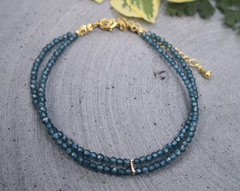 May include: A delicate blue beaded bracelet with gold-colored accents. The bracelet features two strands of small, round beads and a small gold bar with a single gem. The clasp and chain are also gold-colored.