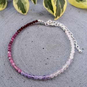 Könnte beinhalten: Ein zartes Armband mit einem Farbverlauf aus Edelsteinperlen. Das Armband besteht aus dunkelroten, rosa und violetten Perlen mit einem silbernen Verschluss.