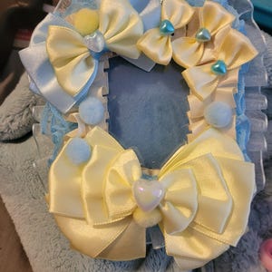 May include: A rectangular photo frame decorated with pale yellow and light blue satin ribbons, ruffled lace trim, small light blue pom-poms, and iridescent heart-shaped charms. The frame features a layered bow design with a central heart accent.