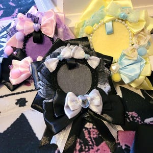 Pode incluir: Uma coleção de laços decorativos em várias cores, incluindo rosa, roxo, amarelo e preto. Cada laço apresenta um elemento central circular, adornado com fitas, renda e enfeites em forma de coração. Alguns laços têm detalhes de pérolas e pompons.