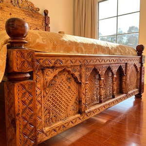 May include: A wooden bed frame featuring intricate hand-carved geometric and floral patterns on the headboard and footboard. The frame has a warm brown finish and is topped with a gold-colored patterned bedspread in a bright room.