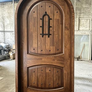 May include: An arched wooden door with a medium brown finish and vertical plank construction. The door features two distinct panels, a black metal decorative grille on the upper section, and multiple dark metal studs along the panel borders.