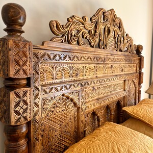 May include: A dark brown wooden headboard featuring intricate hand-carved geometric and floral patterns. The headboard includes a tall, ornate crest with a central pineapple motif and square corner posts topped with spherical finials.